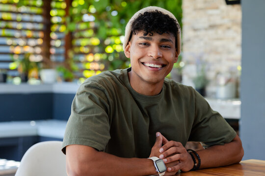 African American man sitting at wooden table on patio, smiling, wearing knit beanie and smartwatch