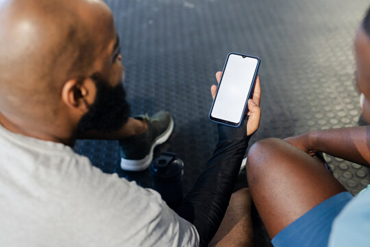 African men sitting on rubber gym floor, one man showing smartphone with black compression sleeve