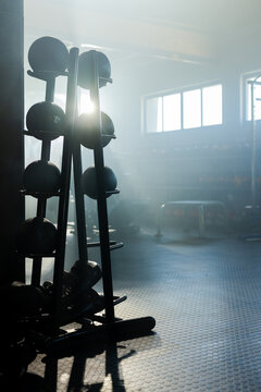 Vertical metal medicine ball rack is holding multiple balls under high windows on rubber floor