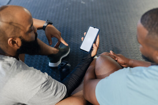 Two African American men sitting on gym floor holding phone pointing at shaker bottle in sportswear