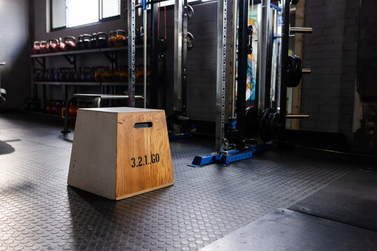Wooden plyo box is resting on rubber coin mats at gym, kettlebells and power rack visible