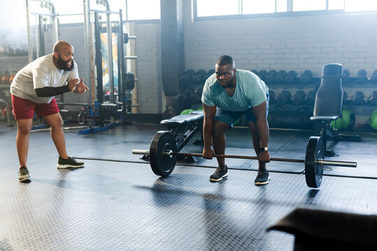 African male trainer and trainee coaching deadlifting in gym wearing sportswear with loaded barbell