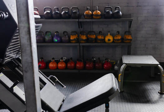 Three-tier metal shelf is holding colorful kettlebells with chipped paint in gym corner