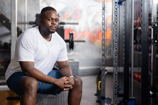 African man sitting on plyo box resting between sets in gym with white tee, barbell, smartwatch