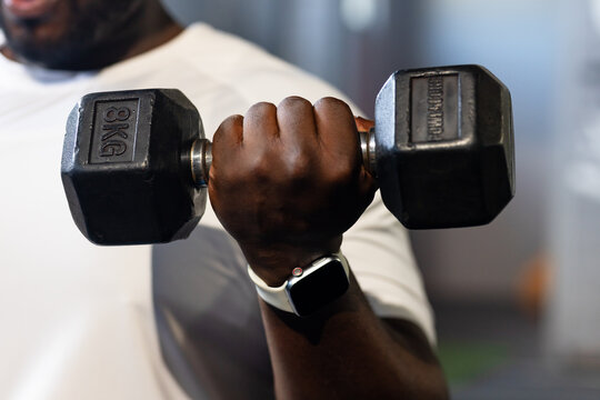 Adult African man gripping 8KG dumbbell, wearing white shirt and smartwatch with white band at gym