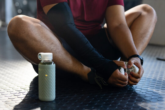 Male athlete is tying white shoe while sitting on gym floor with green bottle nearby