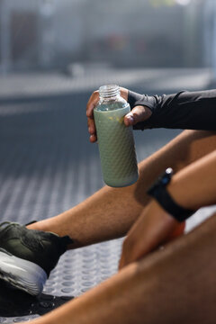 Sitting athlete is holding textured bottle of green liquid on gym floor, showing shoe and watch