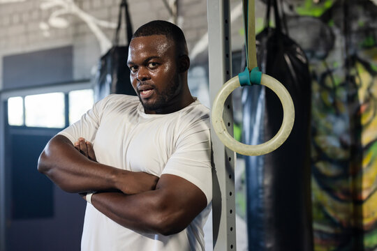 Adult African man leaning against metal support near gym rings in gym, wearing fitted white shirt