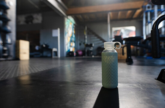 Textured teal water bottle standing on rubber gym mat, casting long shadow, mural visible