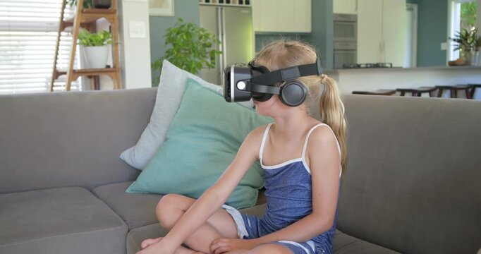 Female preteen wearing VR headset reacting to VR, turning, reaching, smiling on sofa in living room