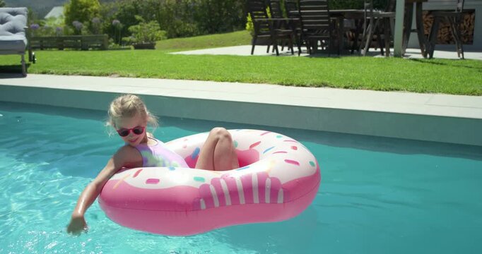 Child girl age 4-6 leaning back then paddling, steering pink donut float at backyard pool edge