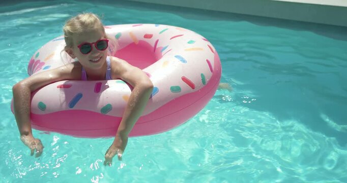 Female child in purple swimsuit smiling paddling and guiding donut float to reach yard pool coping