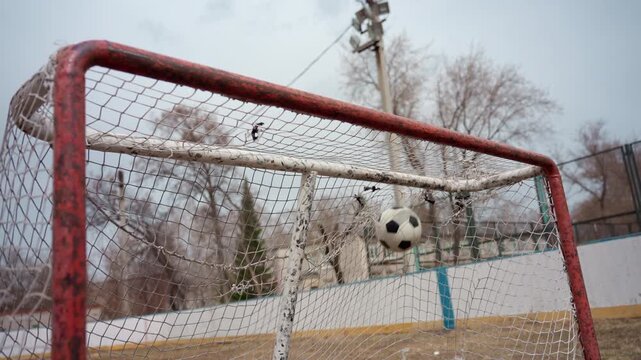 soccer ball hitting net midair. fast moving ball slams into red goalpost net on outdoor rink, blurred background with bare trees and fence, energy and motion captured, splintered paint and gritty