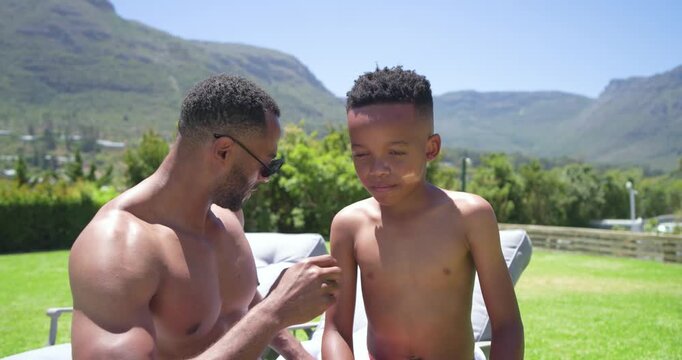 Father and son sitting on lounge chair, pointing at nose and applying sunscreen, protecting skin