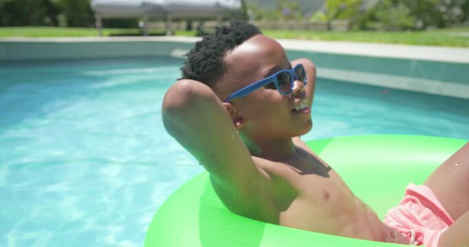 African American youth reclining on green tube in backyard pool squinting from sun relaxing