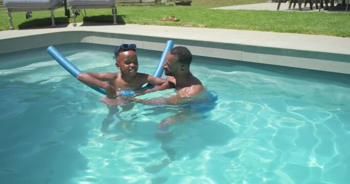 African American father holding son practicing kicks with blue noodle in pink trunks in home pool
