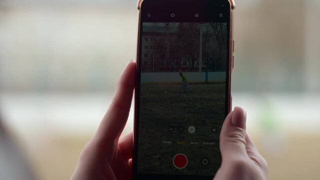 closeup phone recording child on field, hands holding device and red record button visible, blurred window background, gentle indoor light, steady grip capturing distant motion and memory for later