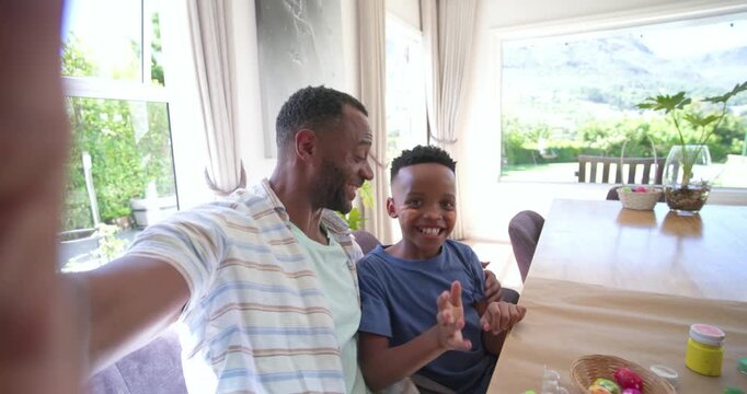 African American father and son holding camera, taking selfies at craft table, painting eggs