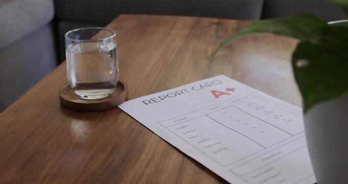 Camera is shifting framing, centering report card and glass on coffee table, revealing letter A+