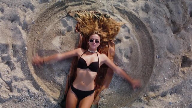 Young Woman Making Snow Angel Shape on Sandy Beach in Black Bikini - Rotating Drone Shot