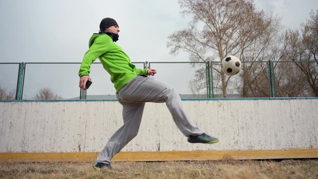 white man practicing soccer tricks outdoors, urban fenced pitch under overcast sky, leafless trees in background, green hoodie and beanie, gray sweatpants, juggling and heading ball, dynamic footwork