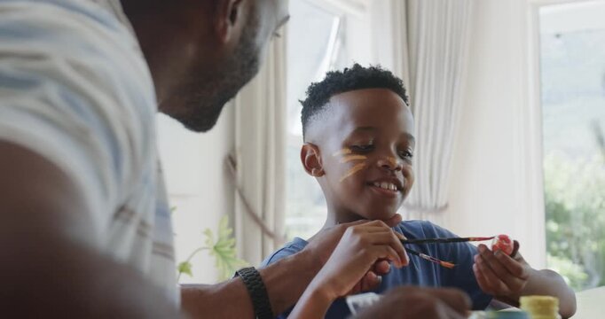 Father and youth son dipping brush, painting face stripes and decorating round toy at home table