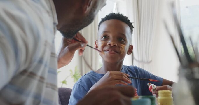 African American father and son at table, father dipping brush, painting son's cheek marks for play