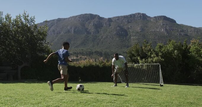 Males mature keeper and youth attacker lunging and dribbling from ball toward goal on backyard lawn