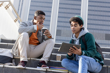 Two college-age males sitting on concrete steps in urban plaza sipping coffee and viewing tablet