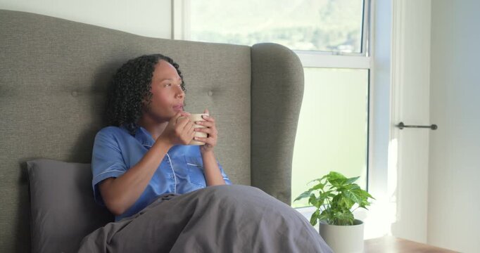 African American woman sitting propped in bed wearing blue pjs holding mug, sunlight urging calm