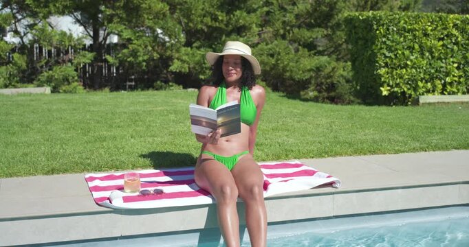 Camera pulling back, Adult African woman in bikini sitting on pool edge reading book dipping feet