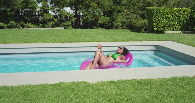 African American woman basking on pink float in yard pool, green suit holding cup, sipping, cooling