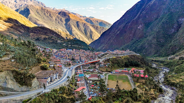 Aerial view of a vibrant town nestled between towering, rugged mountains, where terracotta roofs meet the lush greenery, split by a flowing river, Ollachea, Puno, Peru.