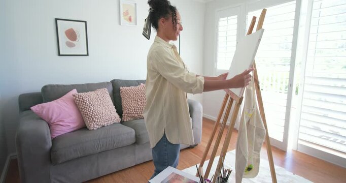African American woman placing canvas on easel, mixing paint at home using ref