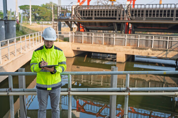 Professional Asian engineer inspecting water quality at a wastewater treatment plant. He is carefully recording maintenance data on a clipboard during an industrial site safety survey.