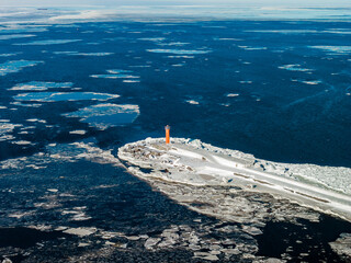 Aerial view of Mangalsala Lighthouse in Riga, Latvia. Bright orange tower stands at icy breakwater as dark water with drifting ice floes extends under crisp daylight. © Aerial Film Studio