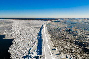 Aerial view of Mangalsala breakwater in Riga, Latvia, as snow lines a jetty between pack ice and pancake ice, with Riga port cranes, icy lighthouse, and clear daylight. © Aerial Film Studio