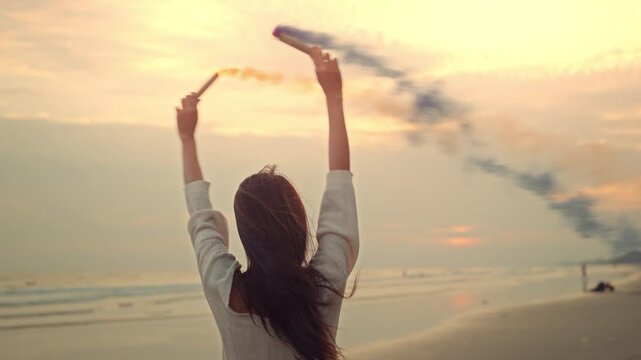 Carefree Asian woman playing with colorful smoke bomb on the beach at sunset, young lady enjoying artistic freedom and celebration during golden hour, concept of creativity, summer vacation