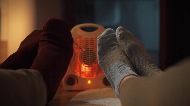 A young couple snuggles beside a warm electric heater on a cold evening, its red glow illuminating their feet, stimulating blood circulation and providing comfort and safety
