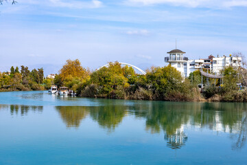 Fototapeta premium Scenic river view with a white lighthouse style tower and a modern bridge reflecting in turquoise water. Autumn landscape with yellow trees and boats under a blue sky in Manavgat, Turkey.