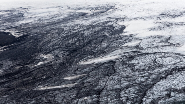 Aerial view of the glacier's icy expanse, marked with dark, winding crevasses against a backdrop of white snow, Iceland.