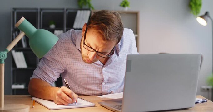 Man with laptop writes notes during analysis. At a home office desk, he organizes documents for audit, verifies report details, and tracks finance figures. Strong concept of financial audit.