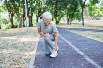 Obraz premium Portrait of a mature middle aged sport woman sitting with a leg injury in pain, after running or exercising in city park outdoors
