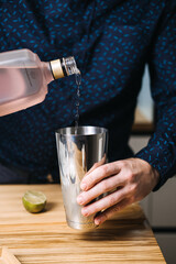 Hand pours clear liquid from frosted pink bottle into stainless steel cocktail shaker. Home bartending, DIY cocktails, hosting at home, mixology kit, social gathering trends, hobbyist bartender.