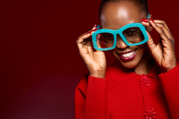 Happy African woman with dark skin wearing bright blue oversized glasses and red sweater smiles joyfully on red background. Concept of fashion, beauty, and cheerful mood.