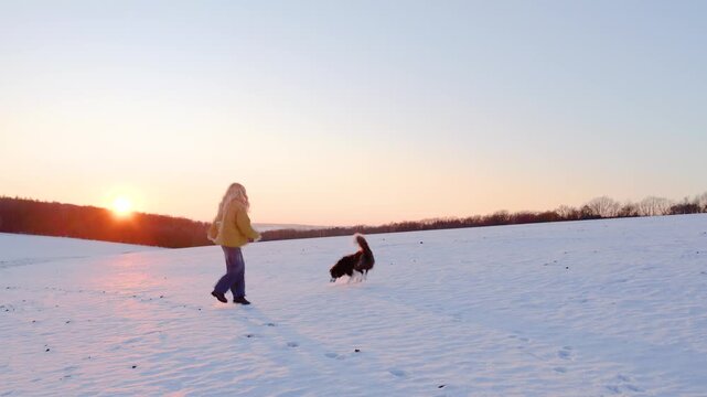 Young woman with long hair playing with her pet border collie dog in the snow, having fun at sunset with happy smile