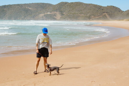 A woman walking a small dog on a leash along an empty beach