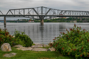 Fototapeta premium Ottawa, Ontario, Canada. View of the wide steel truss Alexandra bridge spanning the Ottawa river, with green shrubs and rocks in the foreground.