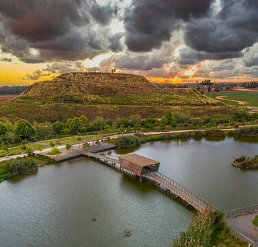 Aerial view of the serene lake reflecting the dramatic sky, with a wooden bridge leading to a covered structure, , Hod Hasharon, Center District, Israel.