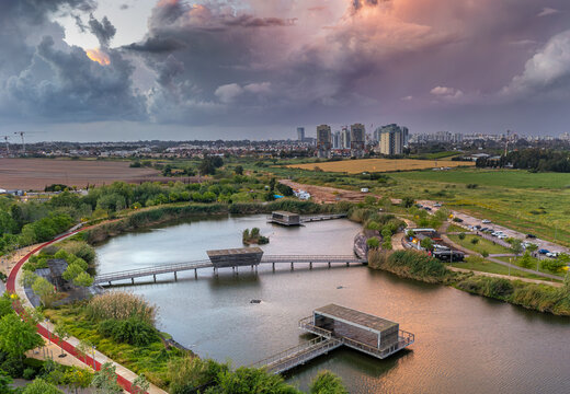 Aerial view of the Hod Hasharon Park's serene lake mirroring the vibrant sunset amidst the urban skyline, Hod Hasharon, Center District, Israel.